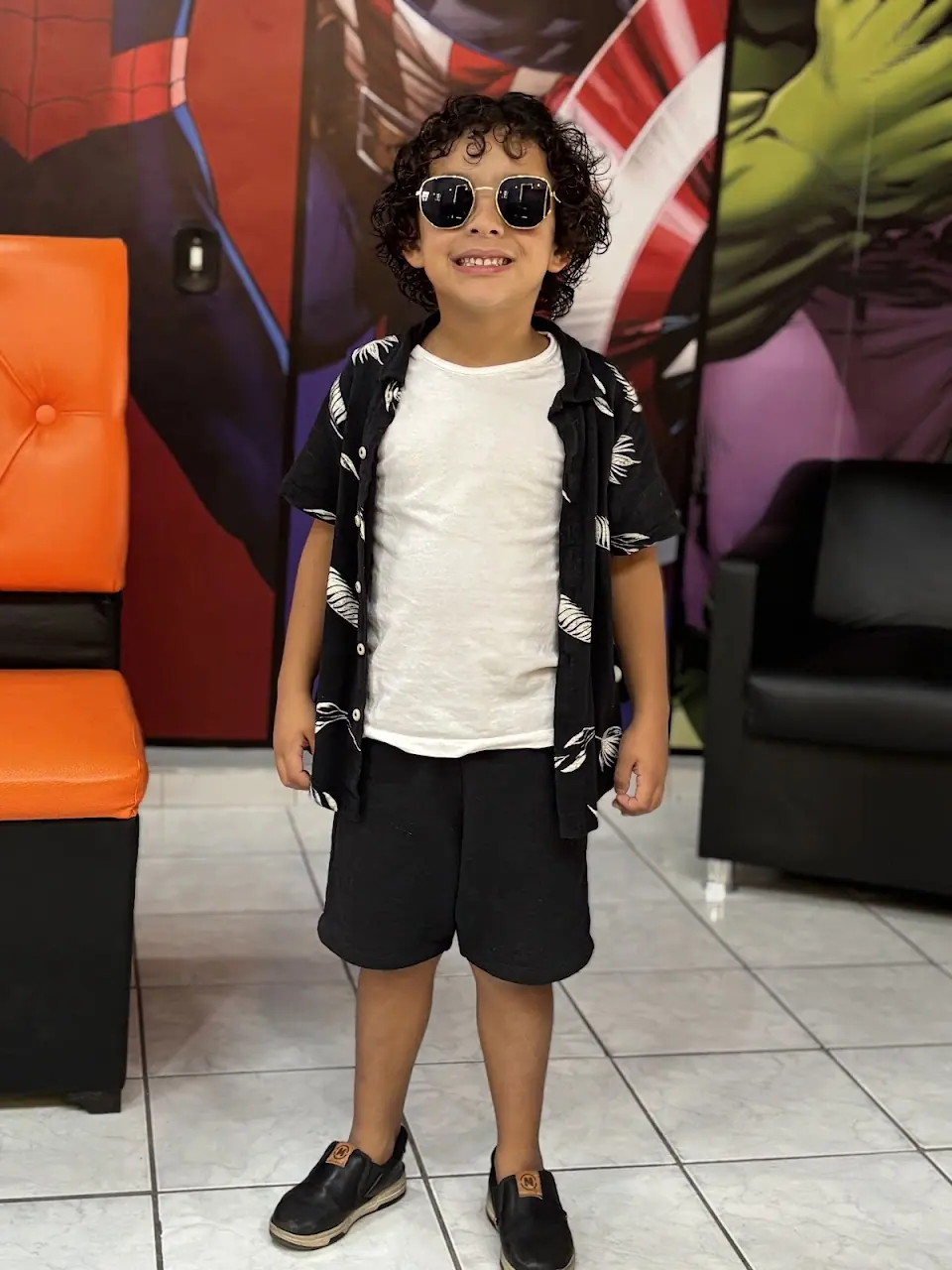 Menino de óculos escuros sorrindo depois do corte, em frente a um painel de super-heróis